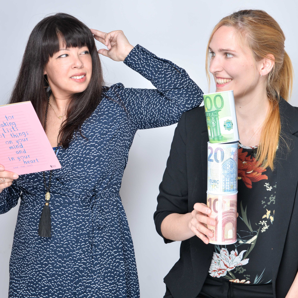 Two women looking at each other, one holding abook, the other holding piggy banks