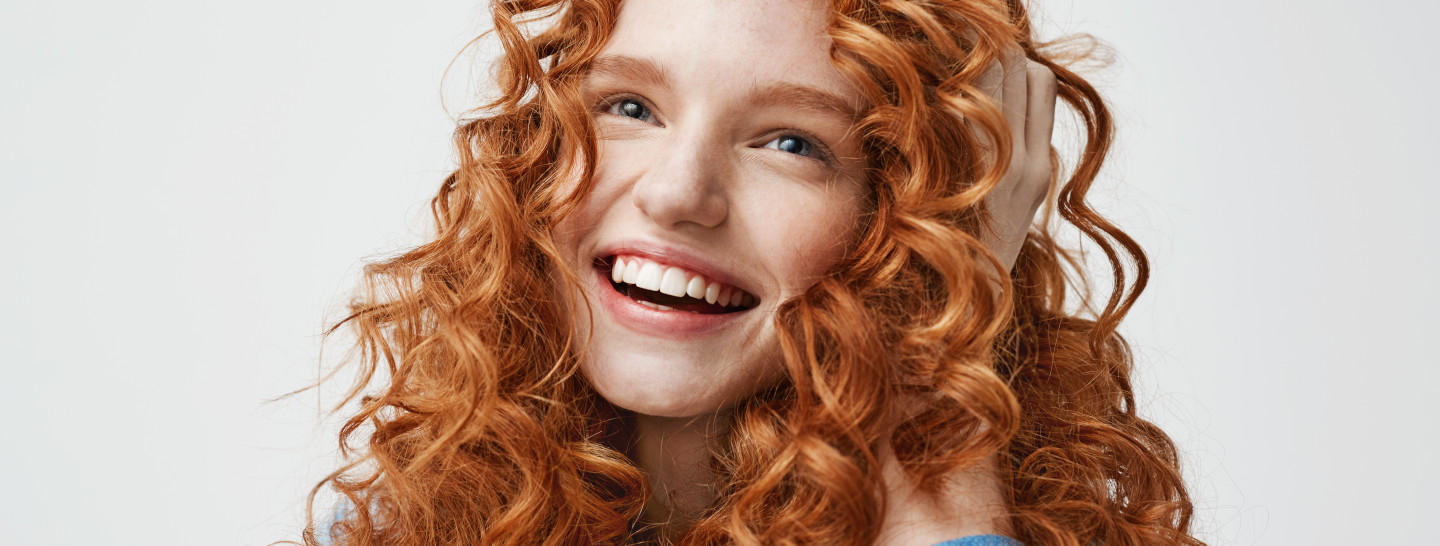 Portrait of cute happy girl laughing touching her curly red hair over white background.